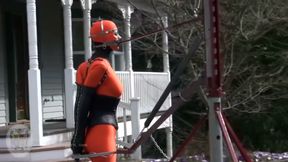 Bound orange latex doll leashed outdoors in front of a porch gets handled rough by her master