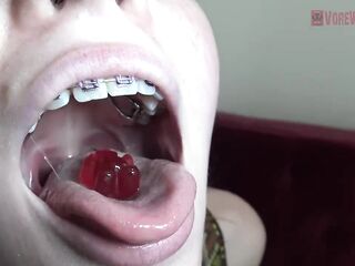 Blue- eyed golden-haired with braces is licking and eating gummy bears in a very hot way